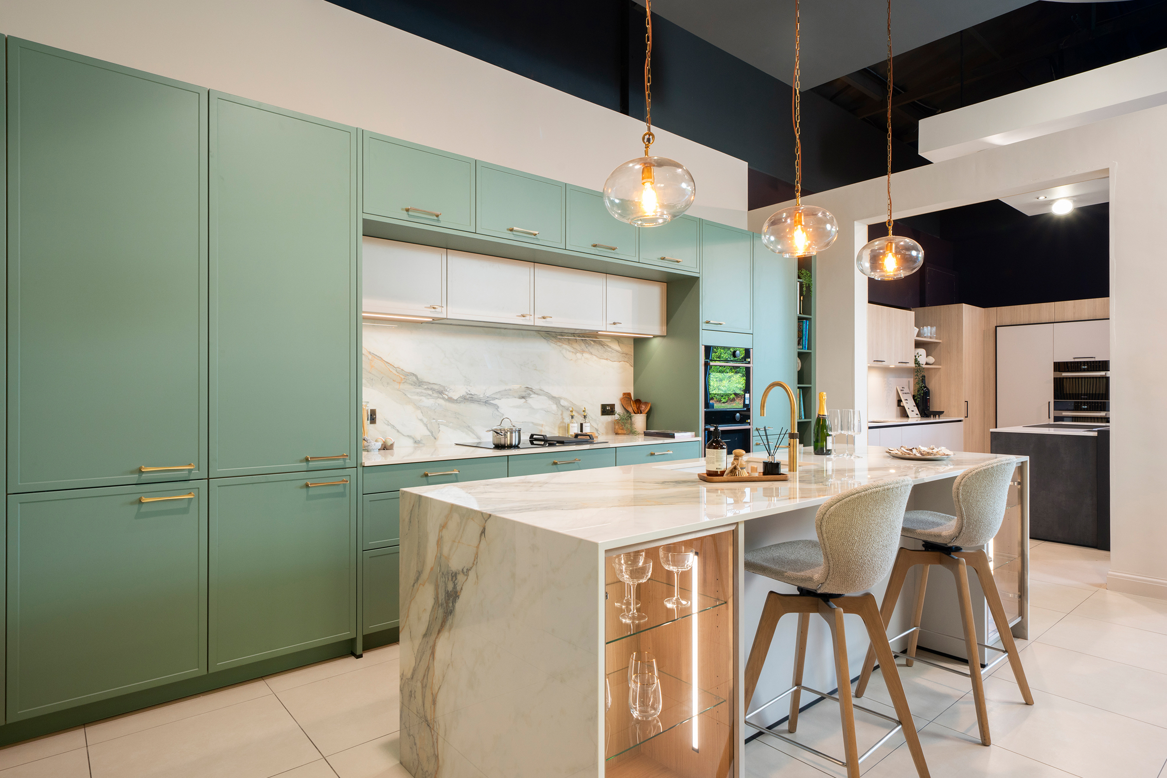 Green slim shaker trade kitchen by KCA featuring a marble-effect quartz island with integrated glass display shelving, brass tap and pendant lighting — showcasing contemporary German engineering and timeless design for developers and interior designers.