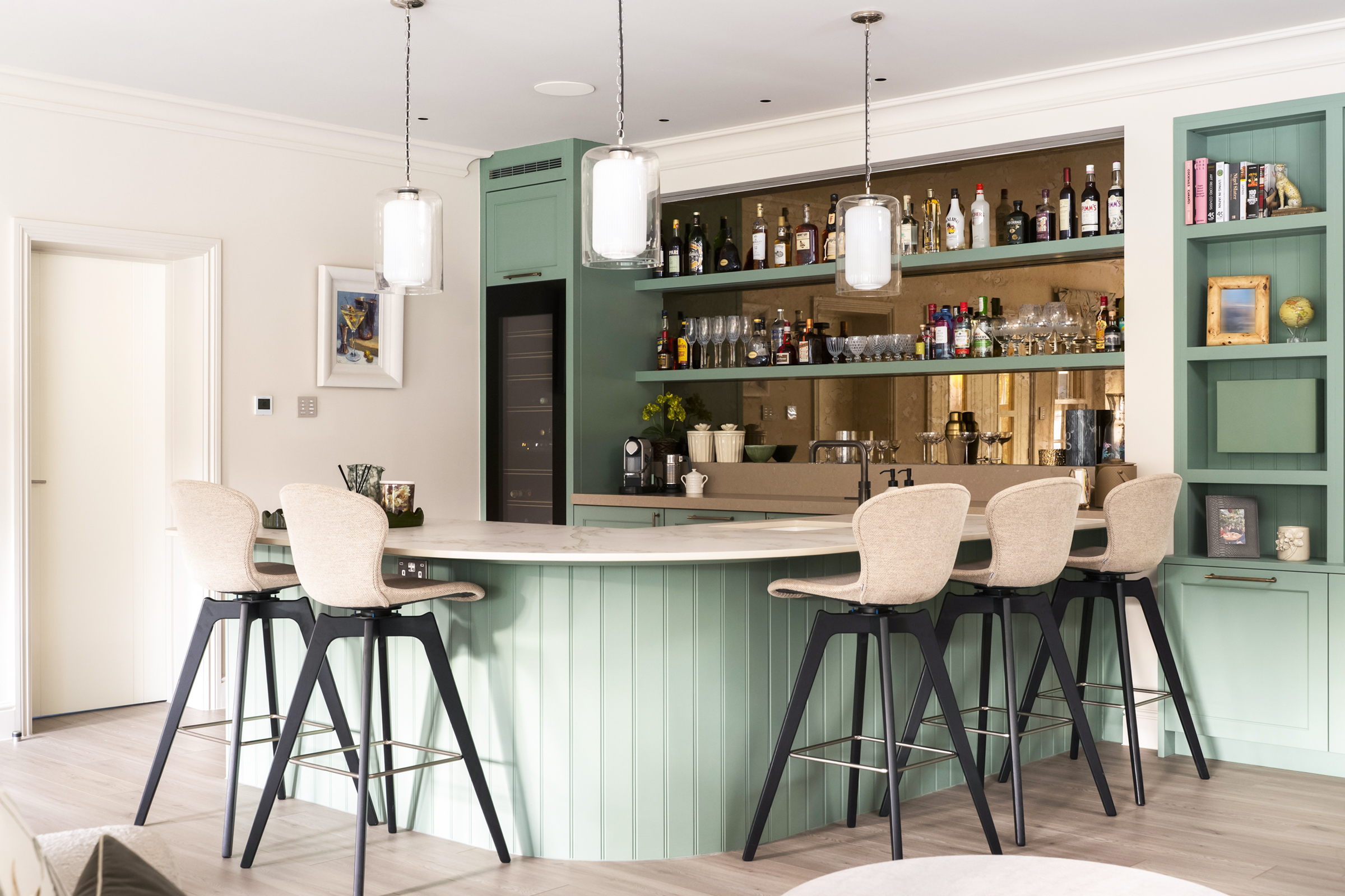 Green curved bar area with tongue and groover paneling, open shelving, bronze mirror and wine cooler.